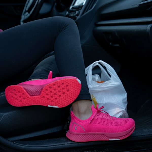 Person wearing bright pink Flux sneakers sitting inside a car with a plastic bag on the floorboard.